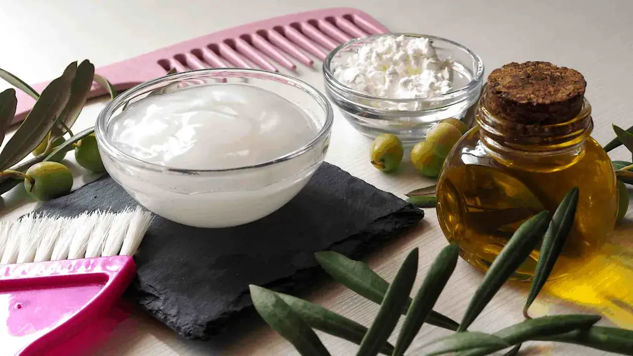 Ingrédients pour un mélange huile cheveux : bol de crème, bol de poudre blanche, olives, flacon d'huile d'olive, peigne rose et brosse.