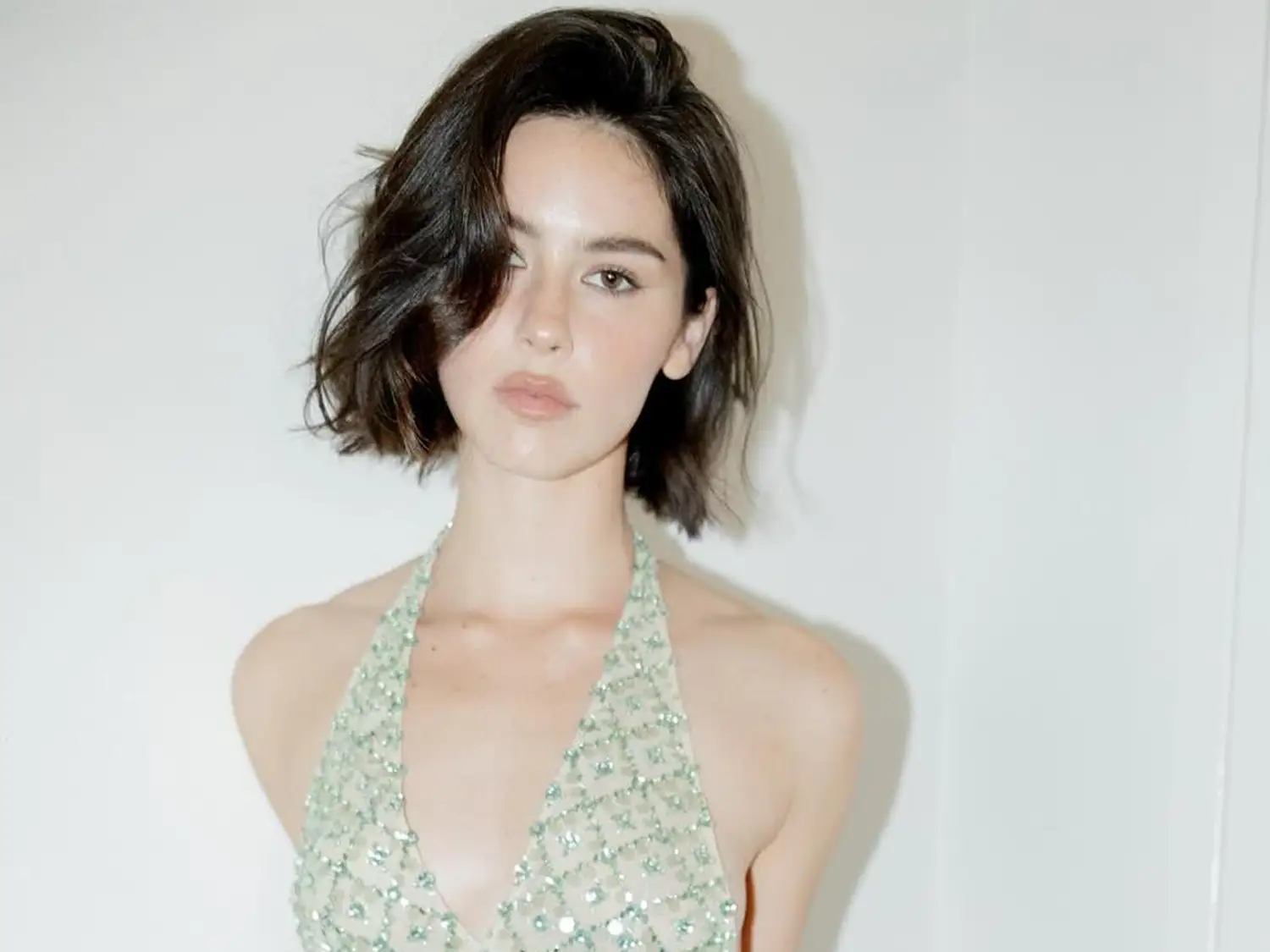 Jeune femme au carr&eacute; flou wavy, portant une robe scintillante, pose devant un mur blanc.