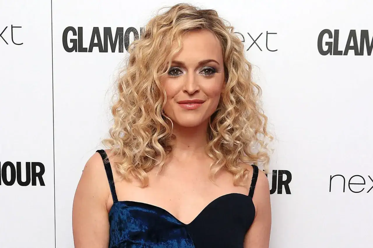 Une femme avec une coiffure année 80, des boucles blondes volumineuses et un maquillage audacieux. Elle porte une robe bleue.
