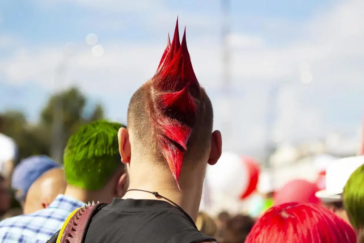 Coiffure festival audacieuse : un crâne rasé avec une crête rouge vif et pointue. D'autres personnes aux cheveux colorés sont visibles en arrière-plan.