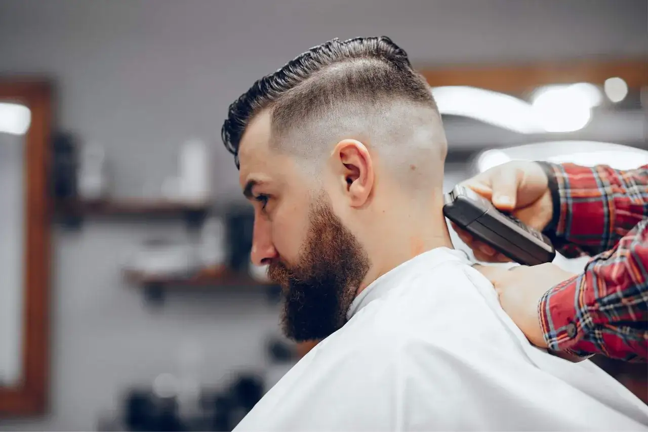 Un homme barbu se fait couper les cheveux, avec une tondeuse qui cr&eacute;e un d&eacute;grad&eacute; net. Le coiffeur, en chemise &agrave; carreaux, travaille le taper long.
