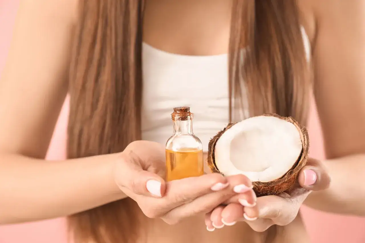 Une femme tient une petite bouteille d'huile de coco et une moitié de noix de coco. L'huile de coco est un remède naturel pour les cheveux blancs.