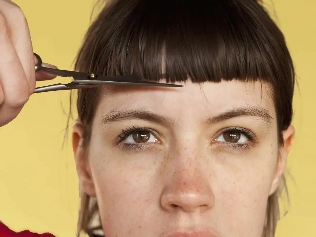 Une main tient des ciseaux pour une coupe rate sur la frange d'une jeune femme aux yeux marron et aux taches de rousseur.