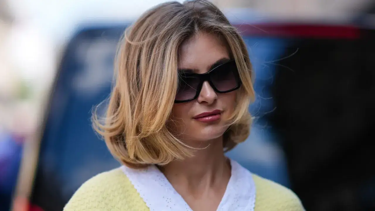 Une femme arbore une coiffure volume femme, avec des mèches blondes ondulées et un pull jaune.