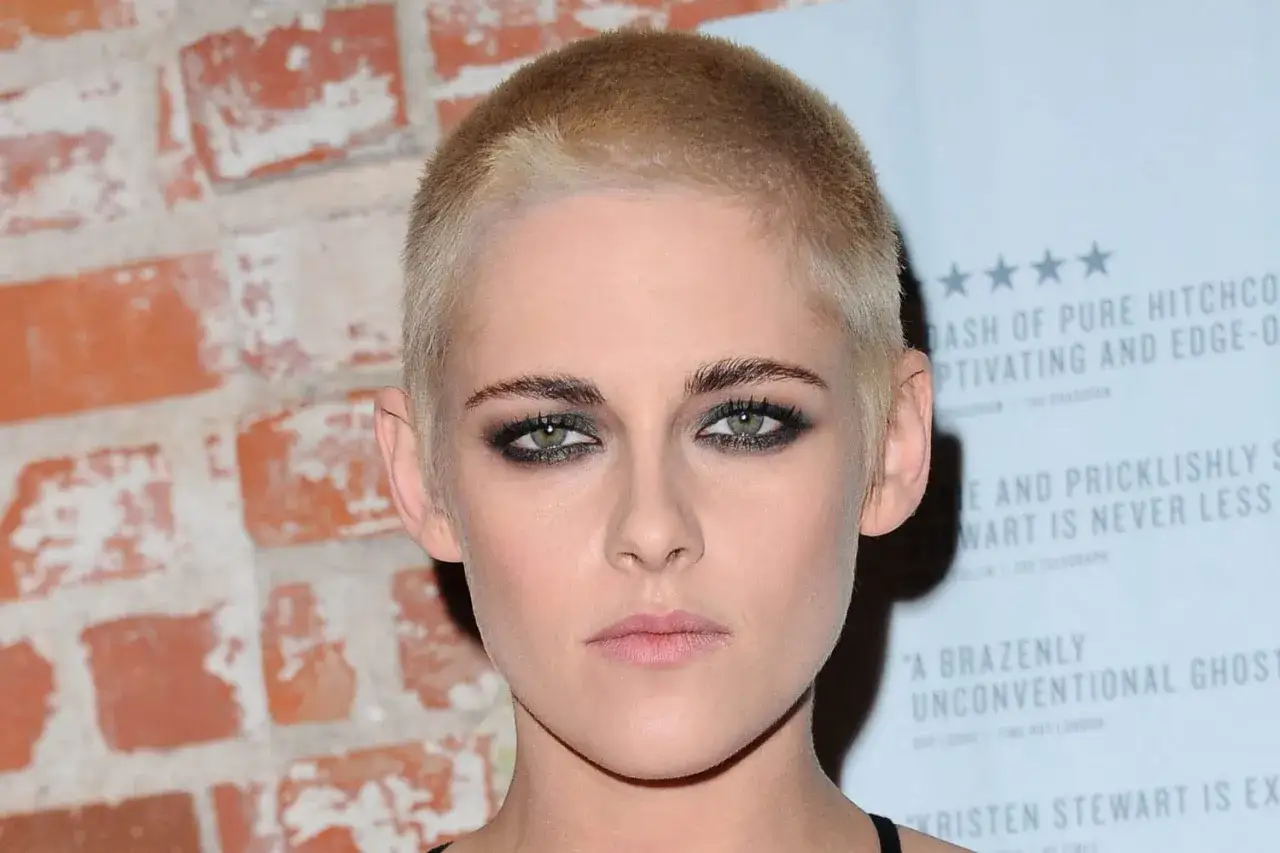 Kristen Stewart, audacieuse avec sa coupe rasee femme, pose devant un mur de briques et un panneau de critiques &eacute;logieuses.