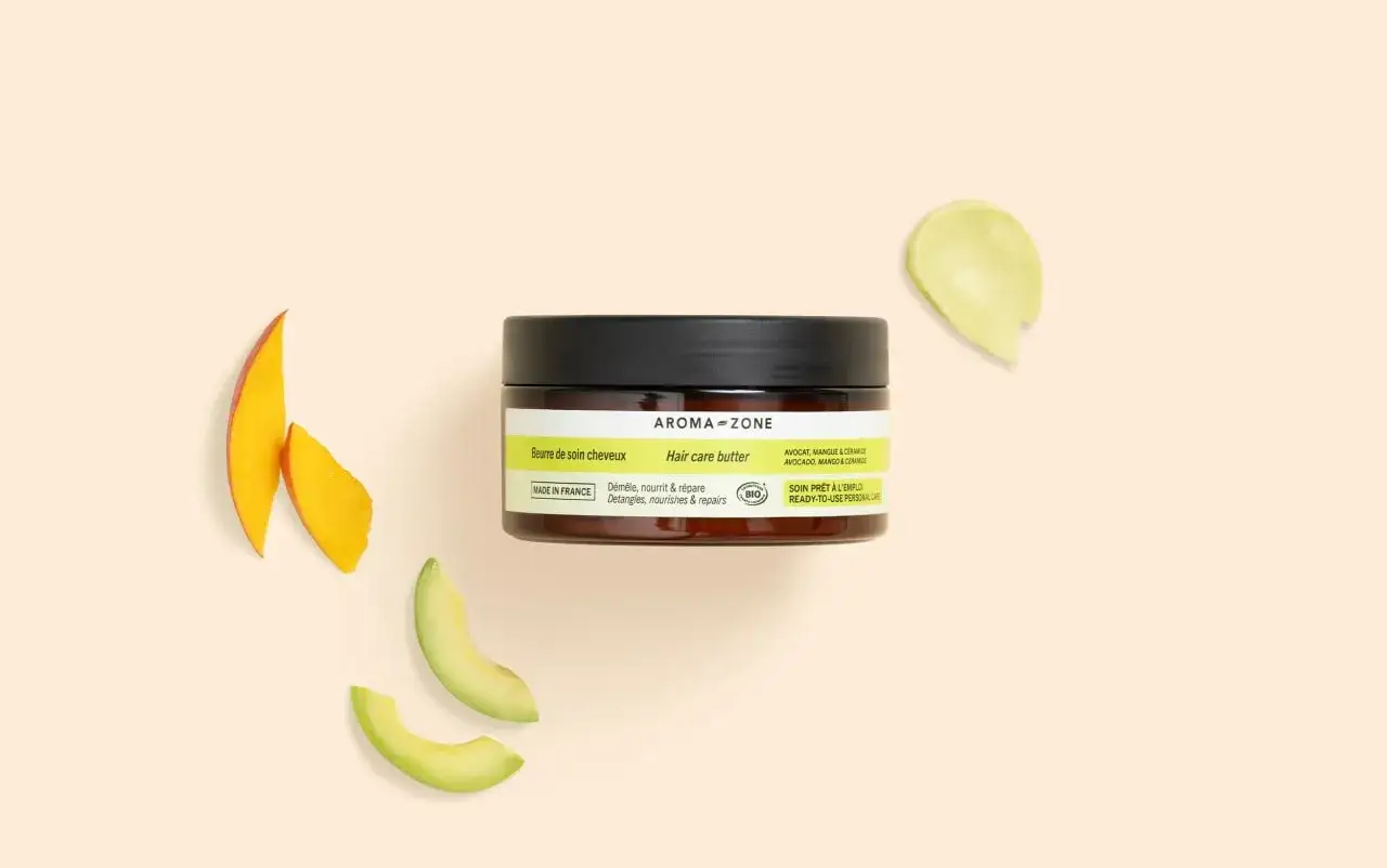 Un pot de beurre de soin cheveux Aroma-Zone, entouré de tranches de mangue et d'avocat. Un soin naturel pour des cheveux nourris.