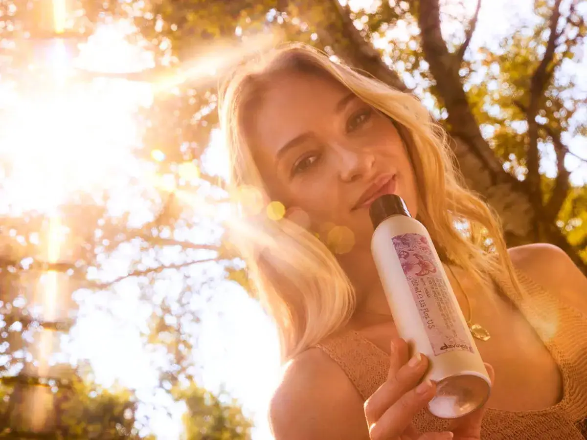 Une femme aux cheveux blonds, style balayage californien, tient un produit capillaire sous le soleil.