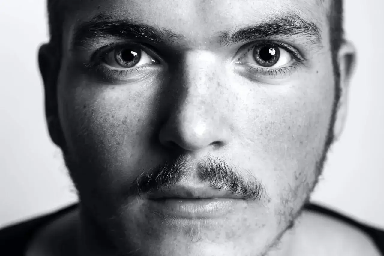 Gros plan sur le visage d'un homme, mettant en valeur ses yeux intenses et un type de moustache subtil, presque naissant.