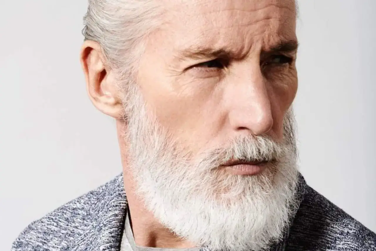 Un homme âgé au regard intense, barbe blanche et coiffure coiffé-décoiffé court, portant un gilet texturé.