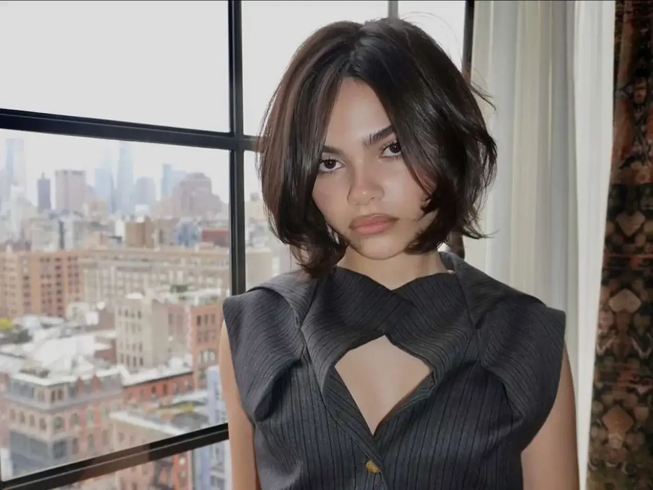 Jeune femme avec un carré court dégradé sur cheveux fins, regardant par la fenêtre avec la ville en arrière-plan.