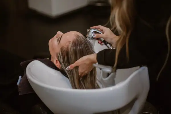Une coiffeuse rince les cheveux blonds d'une cliente. La question "faut il decolorer ses cheveux avant une coloration" est souvent posée avant ce type de soin.
