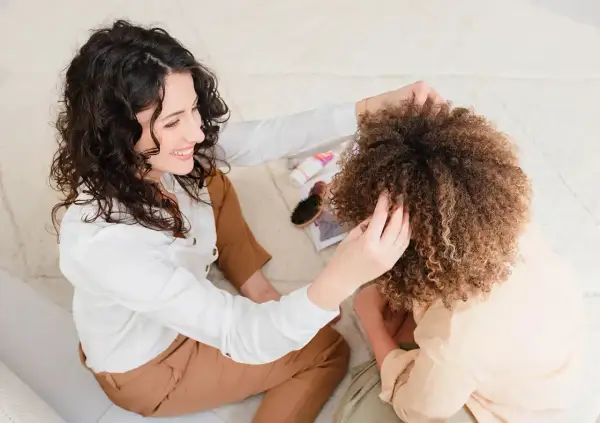 Deux femmes souriantes appliquent un bain d'huile sur des cheveux bouclés, un moment de soin capillaire, que les cheveux soient propres ou sales.