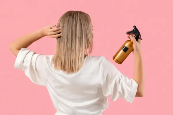 Une femme en peignoir blanc tient un flacon doré, prêt à utiliser du peroxyde d'hydrogène pour éclaircir ses cheveux blonds.