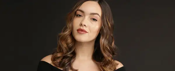 Une femme aux cheveux bruns ondulés, avec des reflets blonds, pose pour une photo. Elle a un maquillage naturel et un sourire subtil. Elle semble avoir réussi à faire un balayage maison.