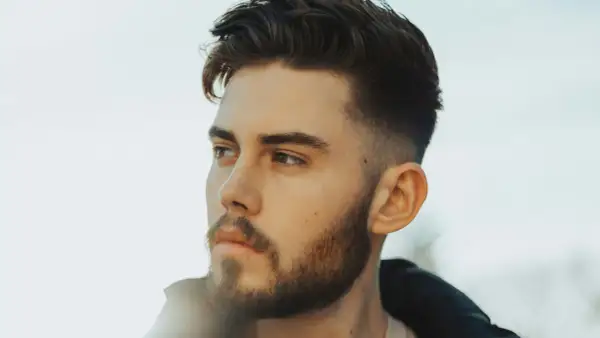 Jeune homme avec une barbe courte et une coupe de cheveux dégradée, style taper fade. Il regarde au loin, le visage éclairé par une lumière douce.
