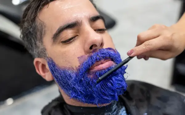Un homme se fait teindre la barbe en bleu vif par une main tenant une brosse.