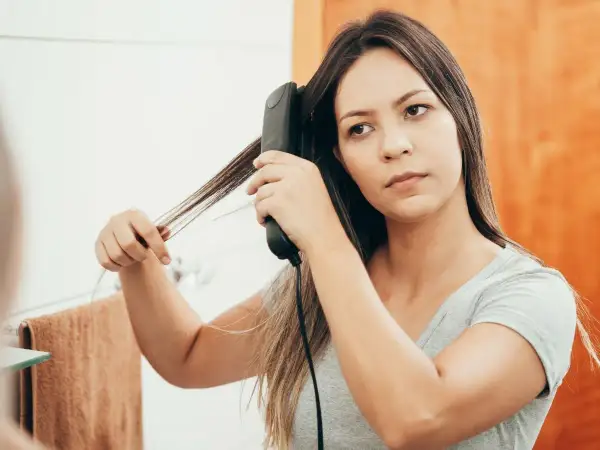 Une jeune femme réalise un lissage brésilien maison devant son miroir, utilisant un fer à lisser pour dompter ses cheveux.