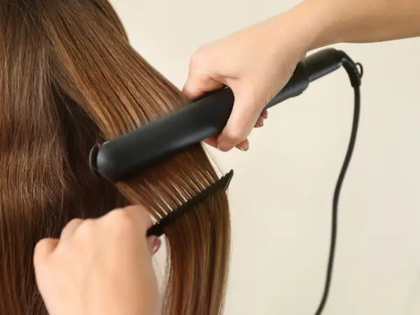 Une main utilise un fer à lisser pour lisser des cheveux bruns brillants, tandis qu'une autre main tient un peigne pour guider la mèche.