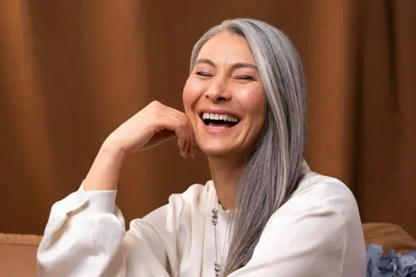 Une femme rit, sa main posée sur sa joue. Ses cheveux gris argenté ont des mèches de couleur subtiles, ajoutant une touche de modernité à son look.