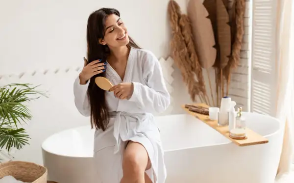 Une femme souriante en peignoir blanc se brosse les cheveux longs et bruns, un geste doux pour nourrir ses cheveux.