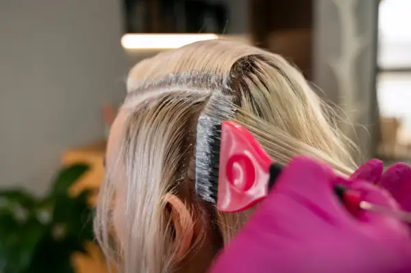 Application d'un produit super éclaircissant ou décoloration sur les racines des cheveux blonds. Une brosse rose et des gants roses sont visibles.