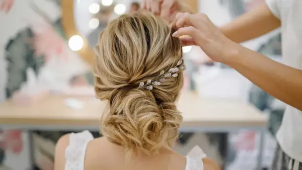 Une mariée se fait coiffer un chignon bas flou, orné d'une barrette scintillante. Les mains d'une coiffeuse ajustent les mèches.