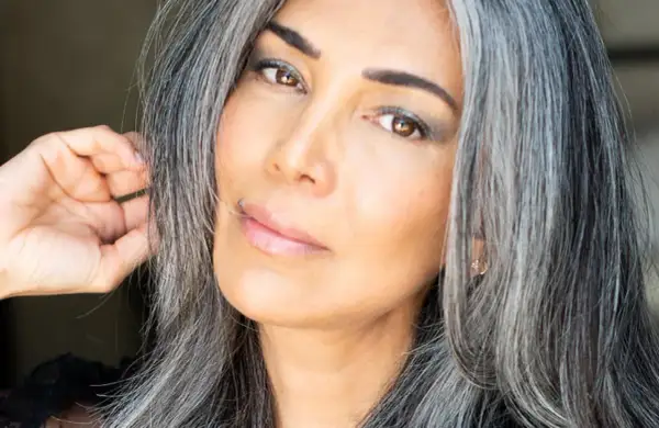 Une femme avec des cheveux gris mi-courts, le regard intense, touche doucement sa chevelure.