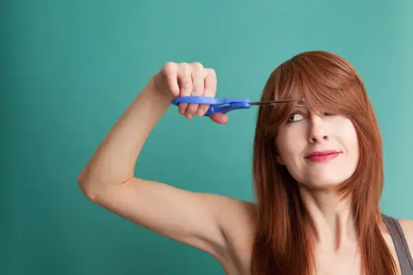 Une femme aux cheveux roux tente de comment couper une frange avec des ciseaux bleus.