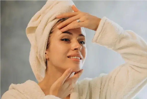 Une femme avec une serviette sur la tête et un peignoir, se touchant le visage. Une astuce pour savoir comment faire une serviette chaude pour les cheveux.
