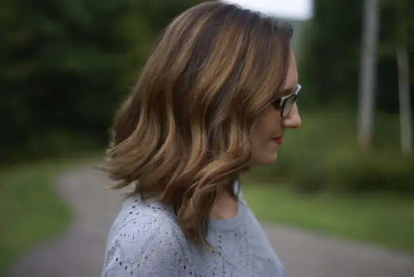 Une femme aux mèches brunes sur cheveux blancs, portant des lunettes, regarde au loin dans un décor verdoyant.