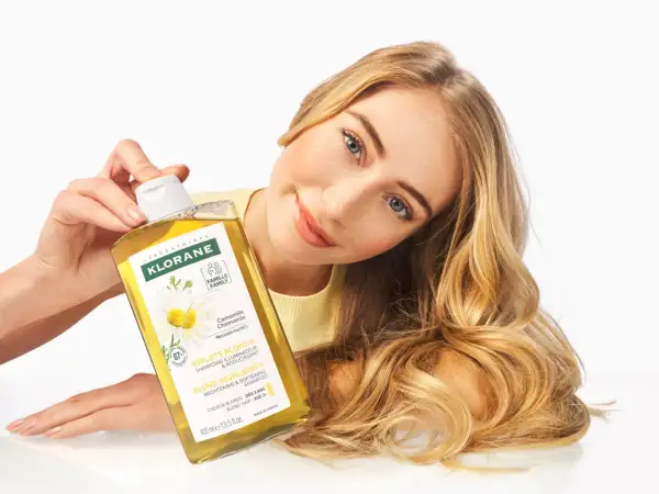 Une femme aux cheveux blonds tient un flacon de shampoing Klorane à la camomille, un produit pour cheveux éclaircissant.